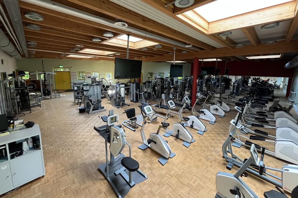 Großer Fitnessraum mit verschiedenen Trainingsgeräten und Holzdecke, helles Licht durch Fenster.