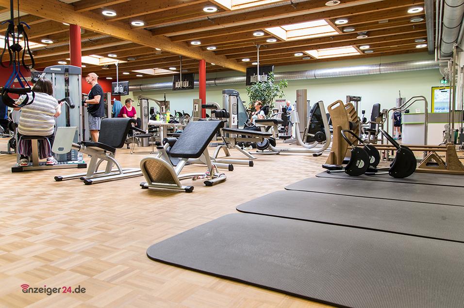 Fitnessstudio mit verschiedenen Ger&auml;ten und Matten, mehrere Personen trainieren im Hintergrund. Holzfu&szlig;boden und Pflanzen sichtbar.
