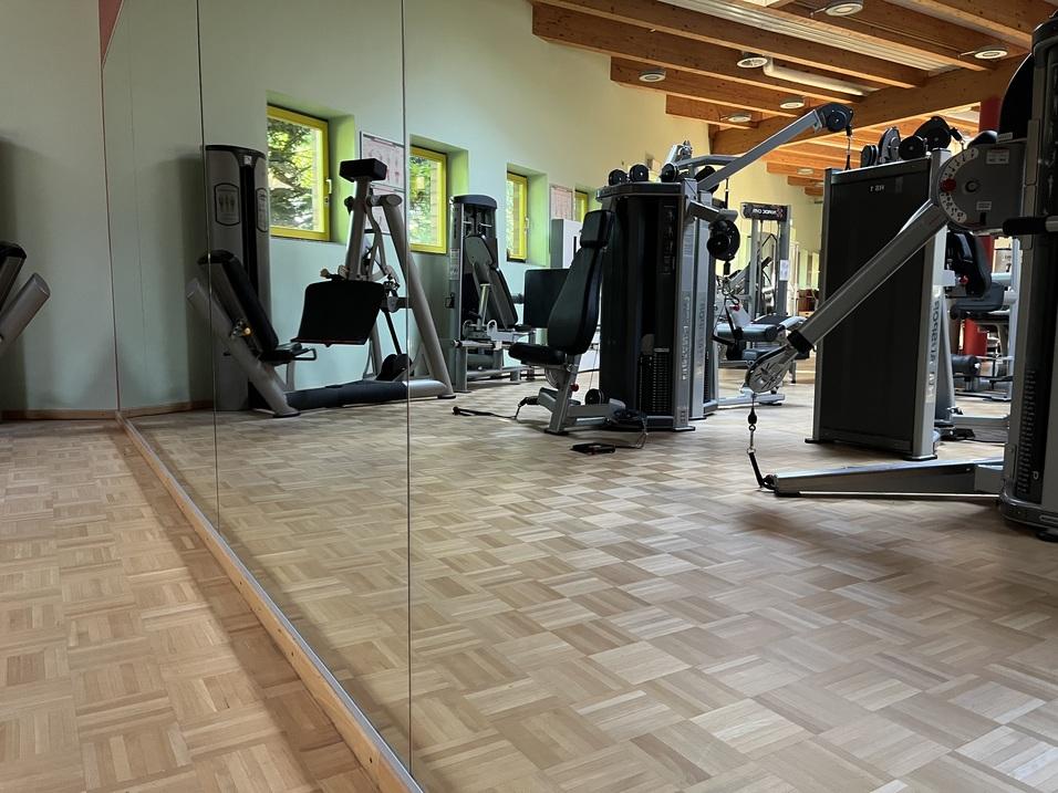 Reflexion im Spiegel eines Fitnessraums mit verschiedenen Trainingsgeräten und Holzfußboden.