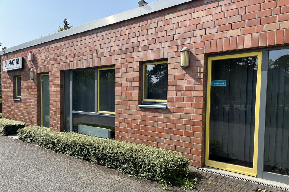 Fassade eines Backsteingeb&auml;udes mit gelben Fenstern und einer gr&uuml;nen Hecke im Vordergrund.