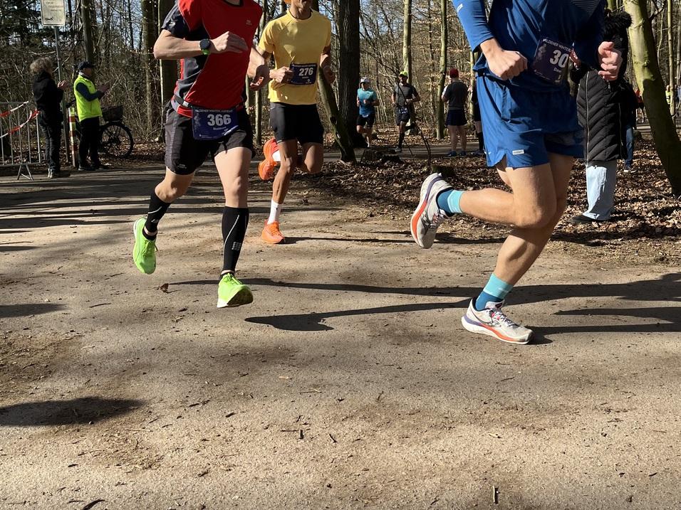 Zwei L&auml;ufer in Sportkleidung beim Wettlauf auf einem Waldweg, umgeben von Zuschauern und weiteren Teilnehmern.