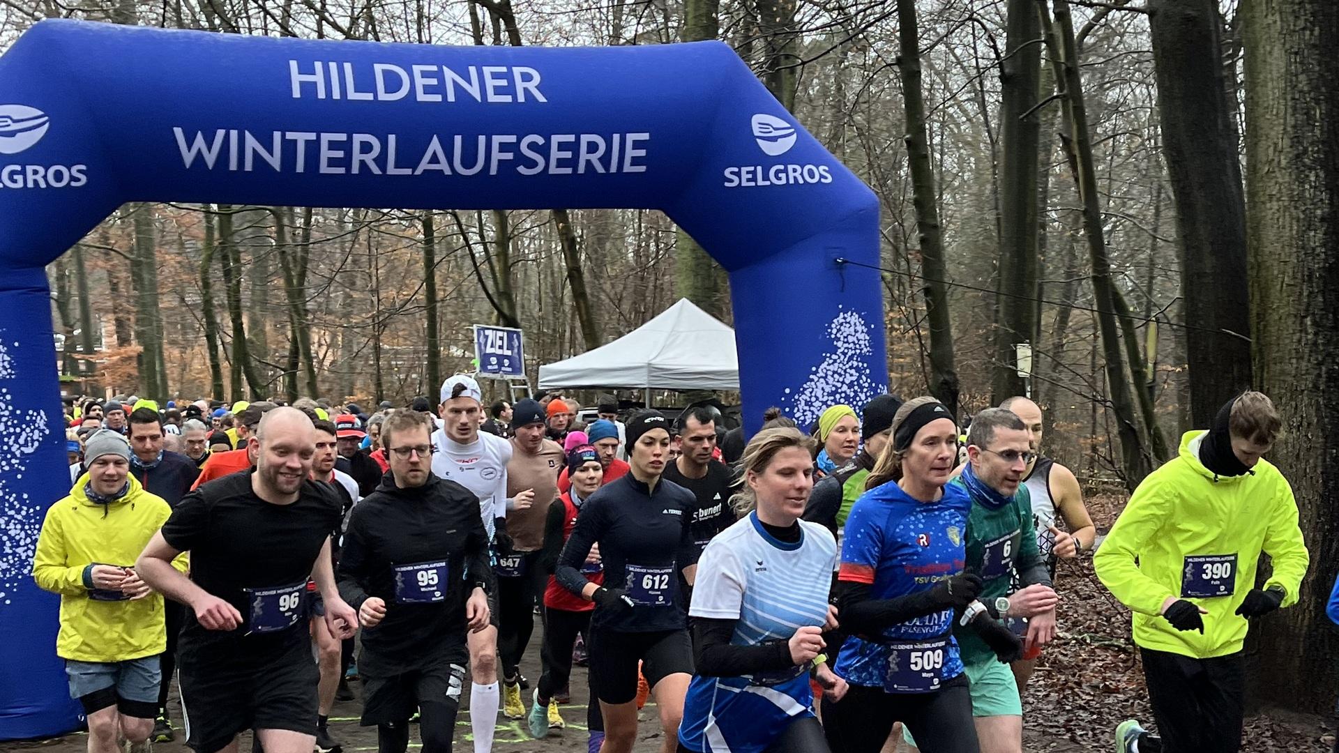 Teilnehmer der Hildener Winterlaufserie starten unter einem blauen Tor in einem bewaldeten Gebiet.