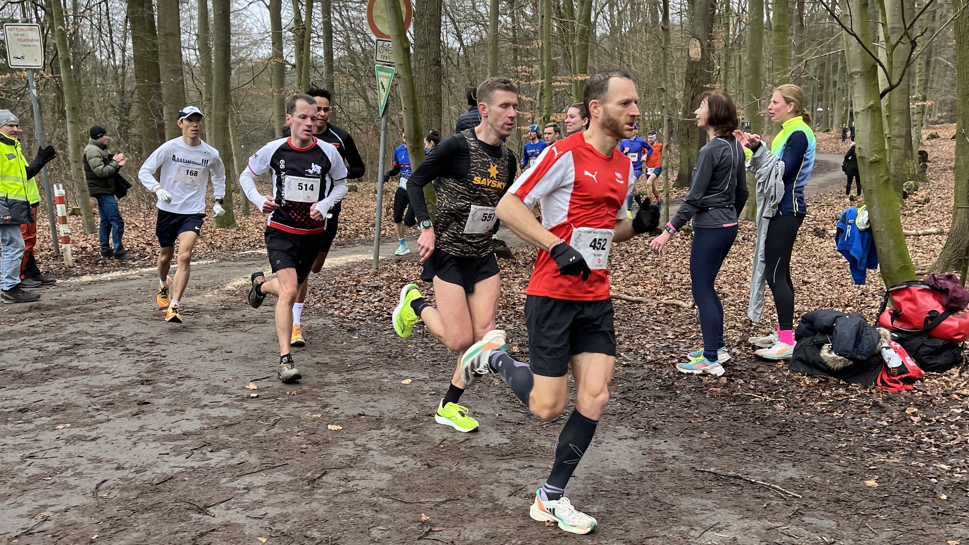 L&auml;ufer in Sportbekleidung joggen auf einem matschigen Weg durch einen Wald, umgeben von Zuschauern.