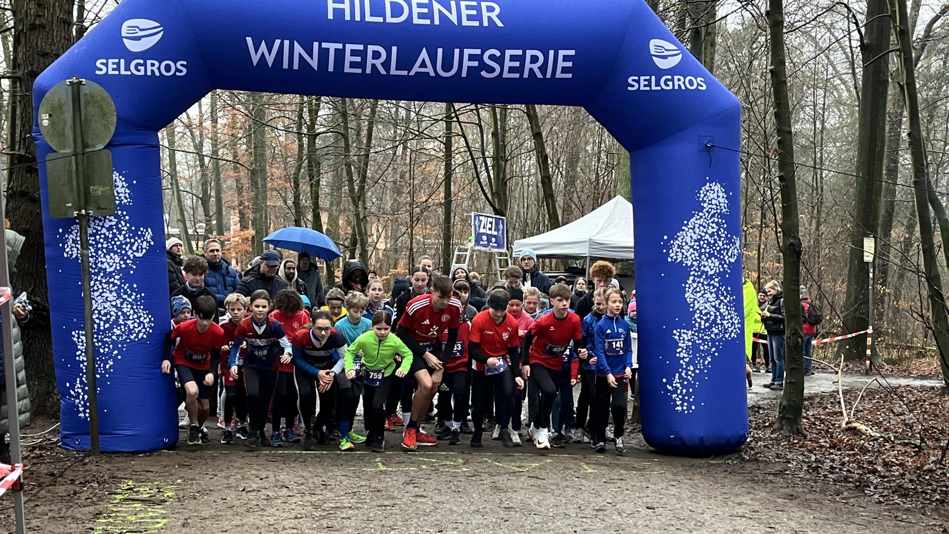 Startgruppe von L&auml;ufern unter einem blauen Bogen mit "Hildener Winterlaufserie" in einem Waldgebiet.