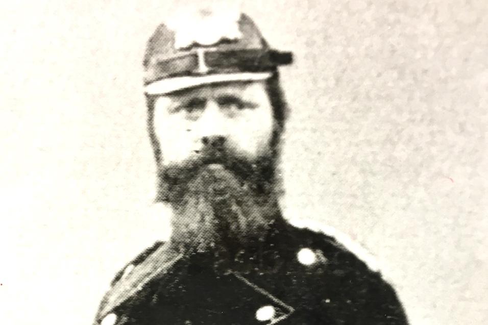 Portrait eines uniformierten Mannes mit Bart und Hut, stehend vor einem neutralen Hintergrund.