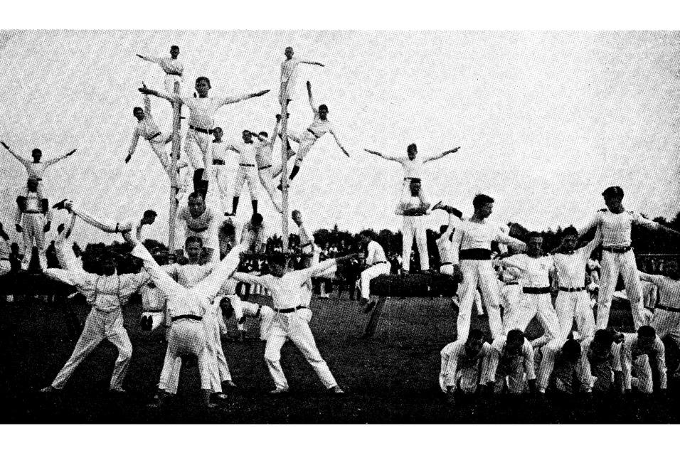 Gruppendarstellung von Turnern in wei&szlig;en Anz&uuml;gen, die akrobatische Figuren auf einem Sportplatz ausf&uuml;hren.