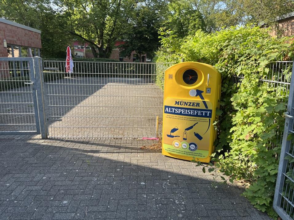 Gelber Altölsammelbehälter steht neben einem Zaun in einem gepflasterten Bereich mit Bäumen im Hintergrund.