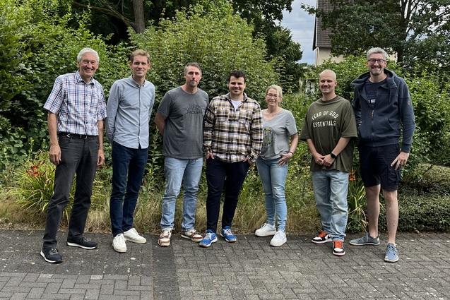 Gruppe von sechs Personen stehen lächelnd auf einem gepflasterten Weg vor einer grünen, bewachsenen Kulisse.