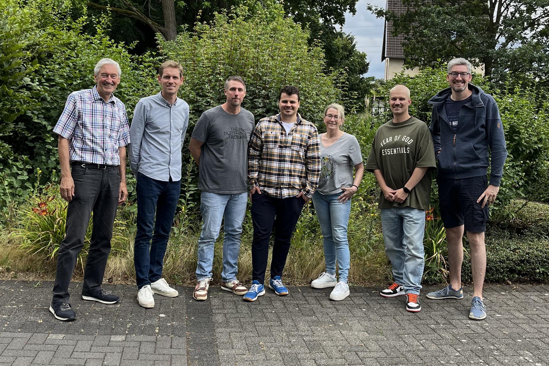 Gruppe von sechs Personen stehen lächelnd auf einem gepflasterten Weg vor einer grünen, bewachsenen Kulisse.