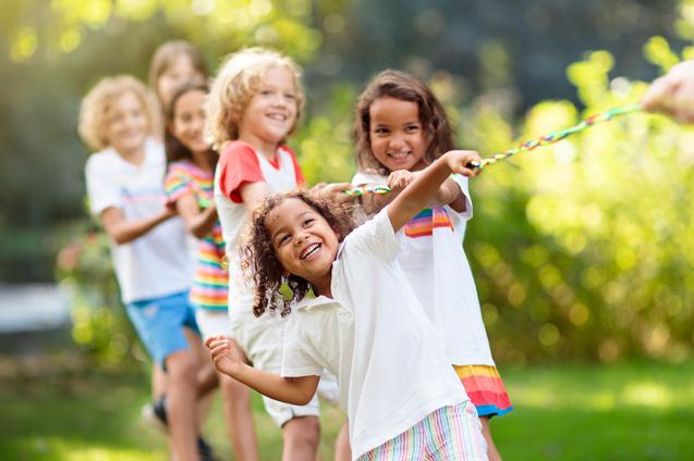 Sechs Kinder ziehen in einem Spiel gemeinsam an einem bunten Seil im Freien, umgeben von Gr&uuml;n.