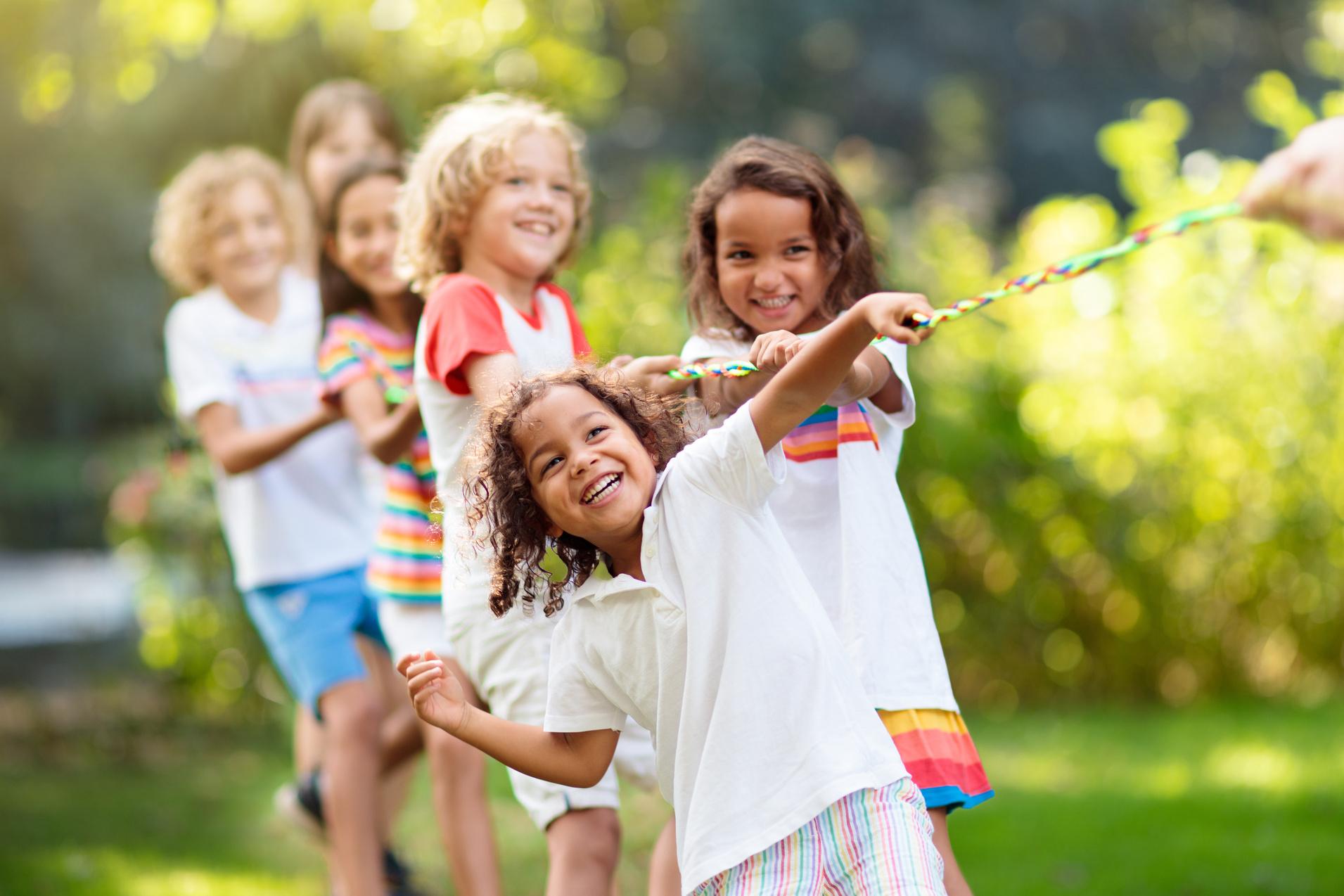Sechs Kinder ziehen in einem Spiel gemeinsam an einem bunten Seil im Freien, umgeben von Grün.