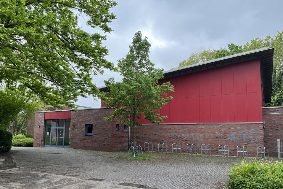 Eingangsbereich eines roten Geb&auml;udes mit gro&szlig;en Fenstern, umgeben von B&auml;umen und einer Pflasterfl&auml;che.
