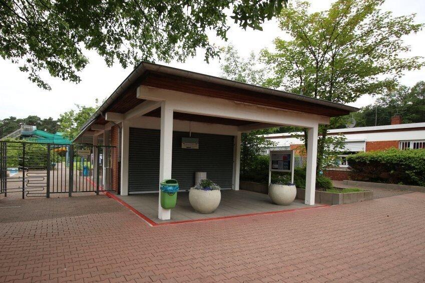 Eingang zu einem Freizeitgel&auml;nde mit &uuml;berdachtem Bereich, K&uuml;belpflanzen und Zugang zu einem Spielplatz im Hintergrund.