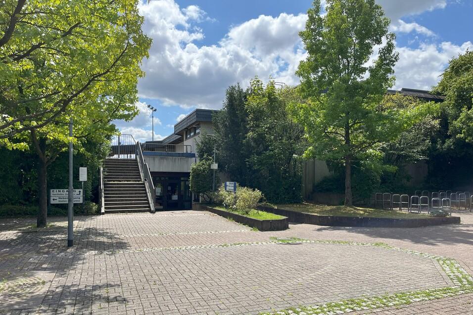 Eingang zu einem modernen Geb&auml;ude mit Treppe, umgeben von B&auml;umen auf einem gepflasterten Platz.