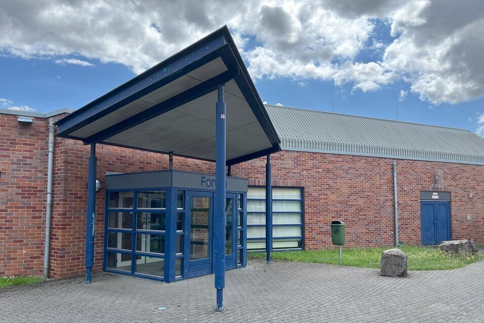 Eingang zu einem roten Backsteingeb&auml;ude mit blauer &Uuml;berdachung und Glasportalen, umgeben von Gr&uuml;nfl&auml;chen.