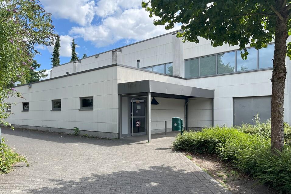 Moderne Geb&auml;udefassade mit gro&szlig;en Fenstern, Eingangsbereich und gr&uuml;ner Abfallcontainer, umgeben von Str&auml;uchern und B&auml;umen.