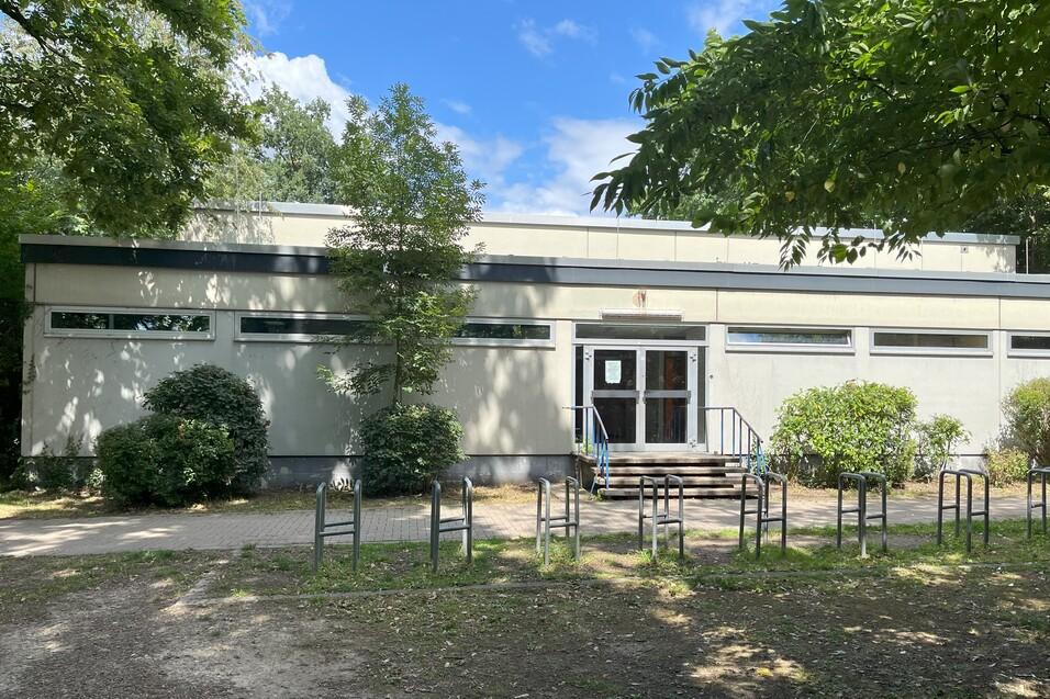 Eingang eines Geb&auml;udes mit Treppe, umgeben von B&auml;umen und Str&auml;uchern, auf einem Weg stehen mehrere St&uuml;hle.