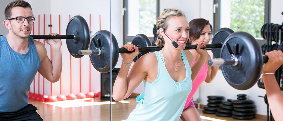 Drei Personen heben Gewichte in einem hellen Fitnessstudio mit großen Spiegeln und Fitnessgeräten im Hintergrund.