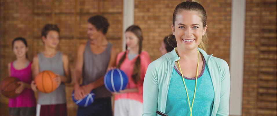 Lächelnde Trainerin mit Stift, im Hintergrund junge Sportler halten Basketballs in einer Sporthalle.