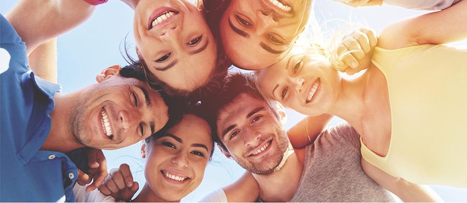 Gruppe junger Menschen, die lächelnd von oben in die Kamera schauen, im Freien unter klarem Himmel.