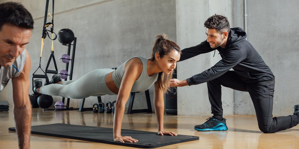 Zwei Personen machen Liegestütze, während ein Trainer einer Frau Unterstützung bietet, in einem modernen Fitnessstudio.