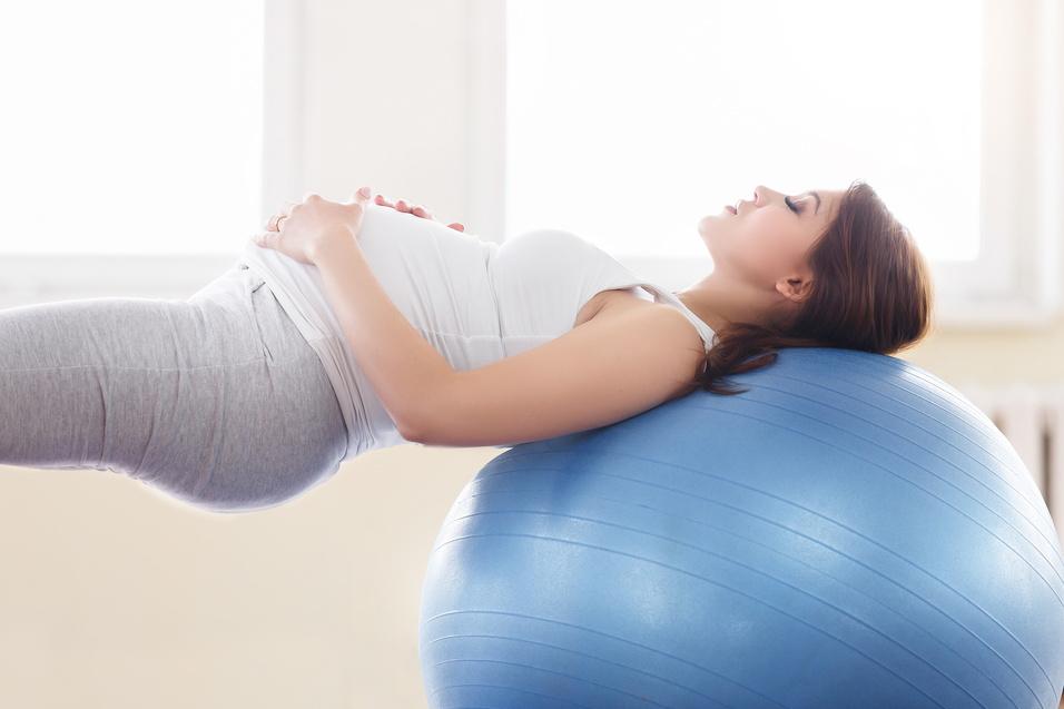 Schwangere Frau liegt entspannt auf einem blauen Gymnastikball in einem hellen Raum.