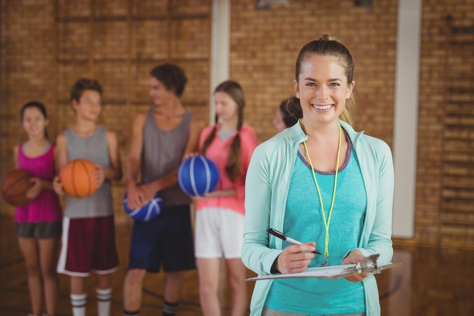 L&auml;chelnde Trainerin mit Clipboard und Stoppuhr, im Hintergrund Jugendliche mit Basketballen in einer Sporthalle.