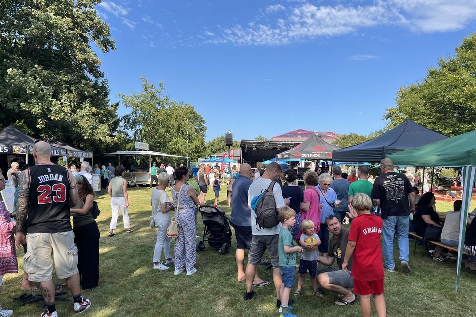 Menschenmenge auf einem Fest im Freien, umgeben von Ständen und Zelten, bei sonnigem Wetter unter blauem Himmel.