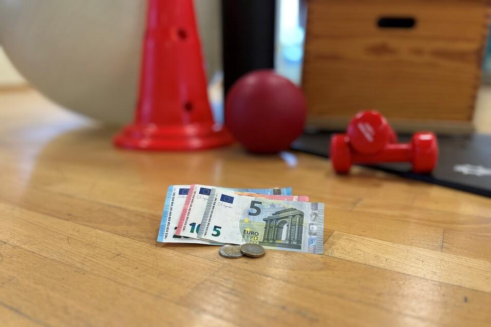 Ein B&uuml;ndel Euro-Scheine und M&uuml;nzen auf einem Holzfu&szlig;boden, umgeben von Trainingsger&auml;ten.