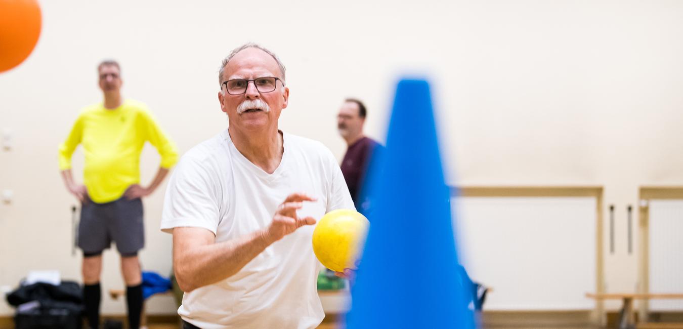 Mann mit Brille wirft einen gelben Ball in einem Sportraum, während andere Teilnehmer im Hintergrund stehen.