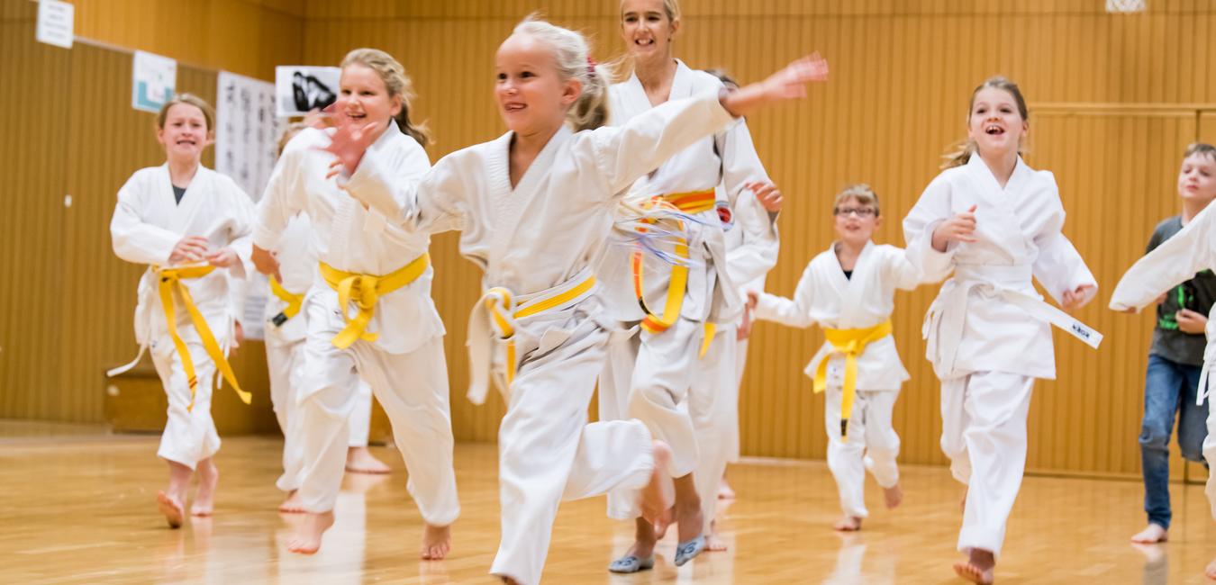 Gruppe von Kindern in weißen Karateanzügen mit gelben Gürteln, die fröhlich in einer Sporthalle herumrennen.