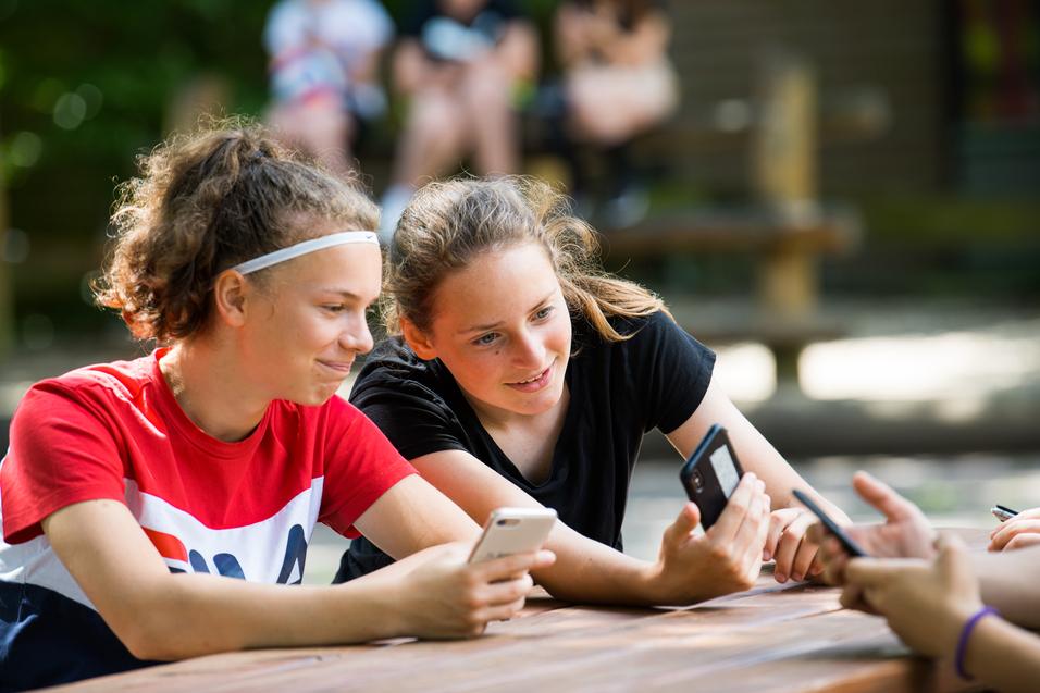 Zwei Mädchen sitzen an einem Tisch, schauen auf ihre Smartphones und lächeln dabei. Im Hintergrund sind weitere Personen sichtbar.