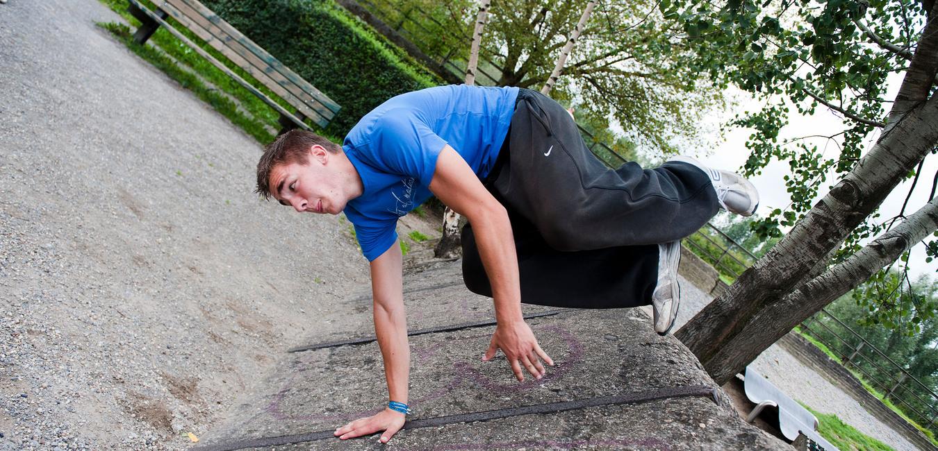 Junger Mann führt eine Parkour-Bewegung auf einer Mauer in einem Park aus, mit Bäumen und einer Bank im Hintergrund.