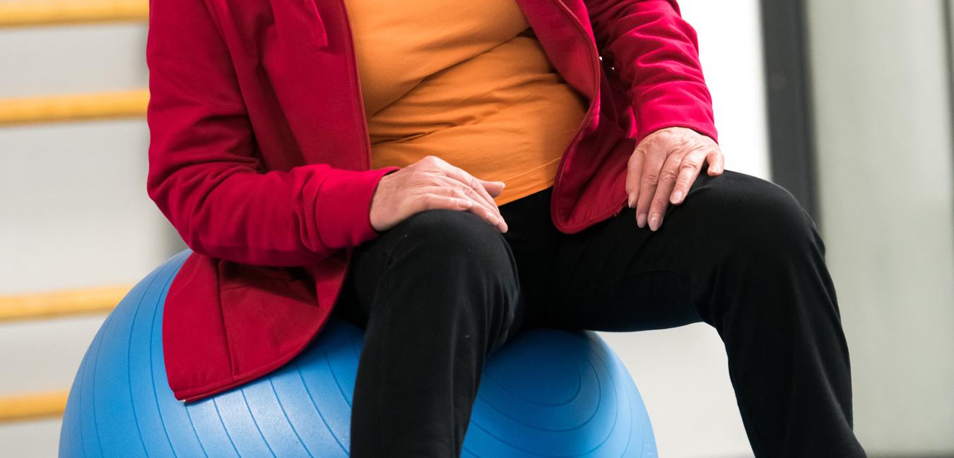 Ältere Frau in roter Jacke sitzt auf einem blauen Gymnastikball in einer Sporthalle.