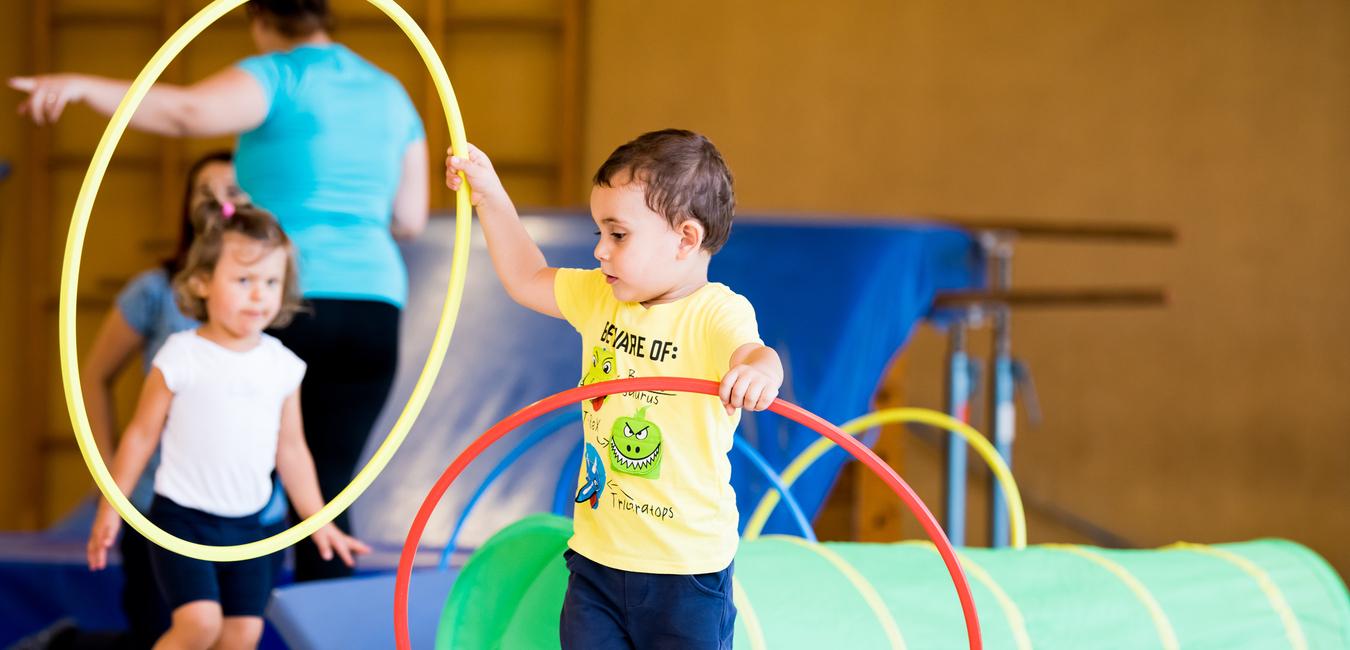 Zwei Kinder spielen in einer Turnhalle mit hula-hoops und einem grünen Tunnel. Ein Kind hält zwei Reifen in der Hand.