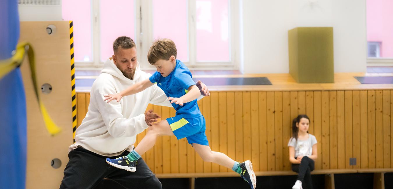 Ein junger Junge springt mit Hilfe eines Trainers über eine Trainingsbank in einer Turnhalle. Ein Mädchen beobachtet.