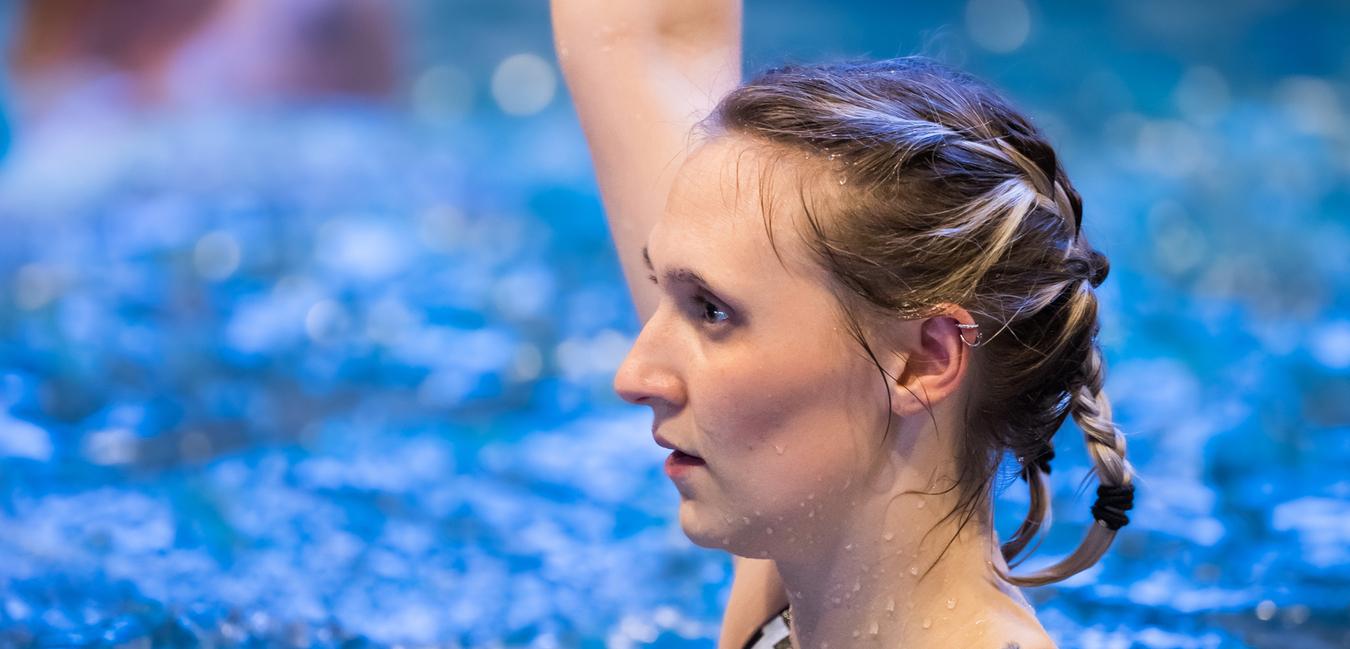 Junge Frau mit geflochtenem Haar hebt ein rotes Aqua-Gym-Gerät über Wasser in einem Schwimmbecken.